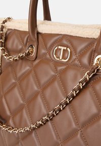 Bolsa em pele castanha com enchimento acolchoado, adornada com cadeado dourado e um forro macio em creme. Possui duas alças e um detalhe de logótipo proeminente.