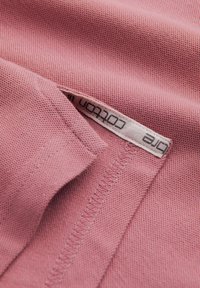 Vaaleanpunainen puuvillakangas, jossa on teksturoitu kudonta, näkyvän ommellun sauman kanssa ja osittain näkyvä etiketti, jossa lukee "Puuvilla".