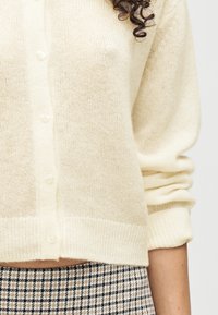 Κρεμ πλεκτή ζακέτα με κουμπιά πάνω από φούστα σε μπεζ, μαύρο και λευκό καρό, εμφανιζόμενη από το μέσο του κορμού μέχρι τη μέση.