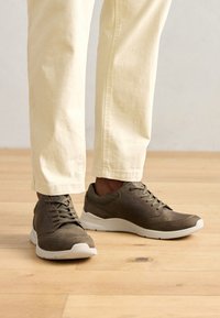 Brown athletic shoes with textured panels, white soles, and laces. Worn with light-colored, slightly cropped pants. Natural wood floor.