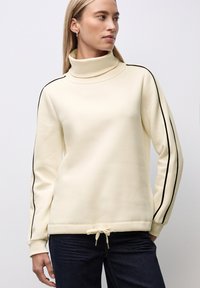 Donna che indossa un maglione a collo alto crema con strisce nere sulle maniche e orlo con coulisse, abbinato a jeans blu scuro.