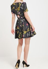Robe noire à motifs floraux avec des manches courtes, taille cintrée et jupe évasée. Portée avec des chaussures à talons hauts noires. Texture de tissu lisse.