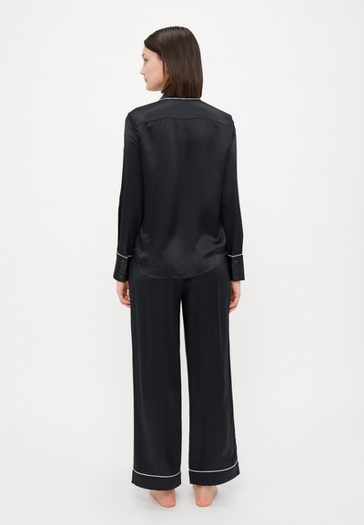 Black satin pajama set featuring long sleeves and wide legs, with contrast piping on cuffs and hem. Soft, smooth texture.