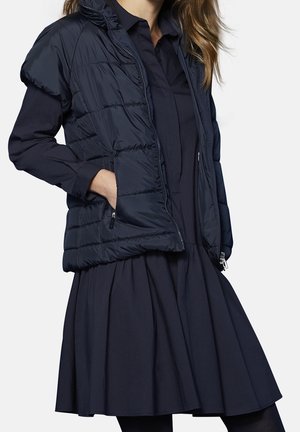 Kobieta w granatowej sukience z długim rękawem i pikowanej bezrękawniku, z ręką w kieszeni kamizelki, stojąca na jednolitym tle.