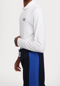 Vit långärmad pikétröja med en svart EA7-logotyp, i kombination med marinblå sweatpants med en blå sidopanel och dragsko i midjan.