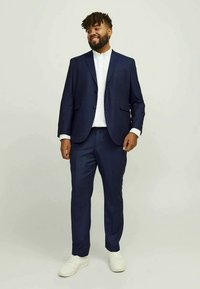 Homem a sorrir, vestindo um fato azul-marinho com uma camisa branca e ténis brancos, em pé contra um fundo claro e simples.