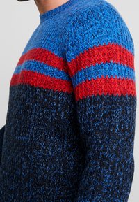 Sweter dzianinowy w niebieskie, czerwone i ciemnonawiwowe paski, z teksturowanym materiałem i okrągłym dekoltem, widoczny w ujęciu z bliska.