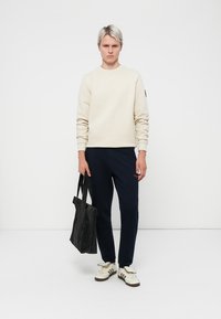 Light beige sweatshirt, navy pants, black tote bag, white and gray sneakers. Simple design, ribbed cuffs, and a small logo on the sleeve.