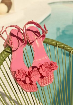 Roosad punutud sandaalid pahkluu rihmadega ja suurte lilledega dekoratsioonidega puhkavad rohelisel traadist toolil basseini lähedal.