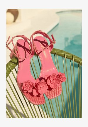 Rózsaszín szövött szandálok bokapánttal és nagy virágos díszítéssel egy zöld huzalos széken pihennek egy medence közelében.