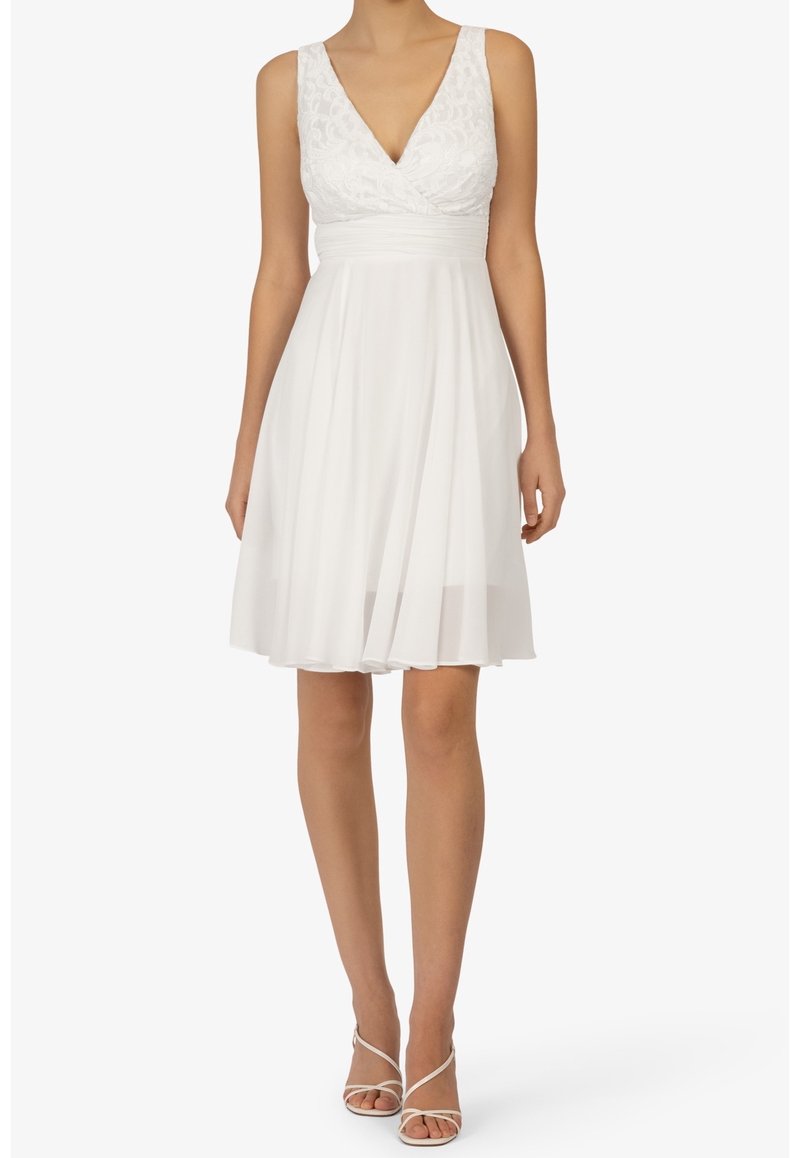 Robe blanche avec un corsage en dentelle et une jupe évasée à mi-genoux. Décolleté en V et bretelles larges, associée à des sandales blanches à lanières.