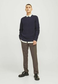 Marineblauer Strickpullover mit V-Ausschnitt, über einem hellgrünen Hemd mit Kragen getragen, kombiniert mit braunen Hosen und schwarzen Schnürschuhen.