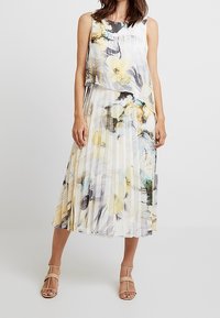 Sukienka z fałdami w kwiatowy wzór w odcieniach żółci i szarości. Lekka, bez rękawów, z warstwowym topem i płynącą sylwetką.