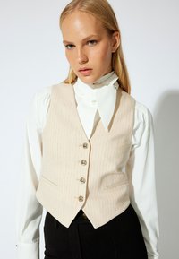 Beige gestreiftes Weste mit taillierter Passform, fünf metallischen Knöpfen, kombiniert mit einer weißen Bluse mit einem Hals-Schal. Schwarze Hosen sichtbar.