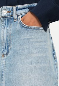Jeans de denim azul claro com cintura alta, apresentando costuras laranjas, um bolso frontal e fecho de botão metálico. A textura parece macia.