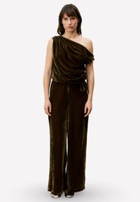 Donkergroene fluwelen jumpsuit met één schouder, gedrapeerde stof, wijd uitlopende pijpen en een getailleerde taille met strikdetail. Gladde textuur, formele stijl.