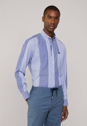 Camicia a righe blu e bianche con righe verticali e orizzontali a contrasto, indossata con pantaloni azzurri con coulisse.