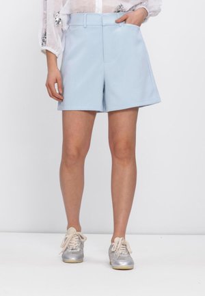 Vrouw draagt lichtblauwe op maat gemaakte shorts met riemlussen, een witte doorschijnende blouse met pailletten en zilveren vetersneakers, staand tegen een witte achtergrond.