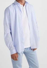 Ljusblå och vitrandig långärmad skjorta, knäppning framtill, med en ficka; bärs över en vit pikétröja och matchas med ljusblå jeans.