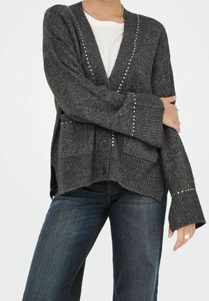 Personne portant un cardigan en maille gris foncé avec des clous argentés, une chemise blanche et un jean bleu foncé, debout avec un bras croisé sur la poitrine.