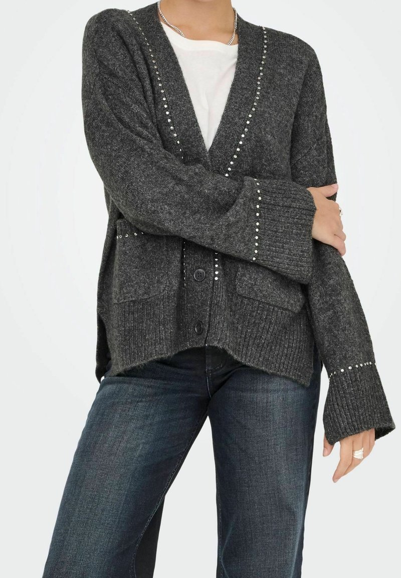 Personne portant un cardigan en maille gris foncé avec des clous argentés, une chemise blanche et un jean bleu foncé, debout avec un bras croisé sur la poitrine.