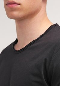 Młoda osoba ubrana w gładką czarną koszulkę z okrągłym dekoltem, pokazująca dolną część twarzy, szyję i górną część klatki piersiowej na neutralnym tle.