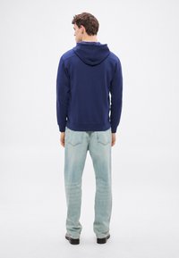 Tengerészkék kapucnis pulóver bordázott mandzsettákkal és szegéllyel, világoskék farmernadrággal párosítva. A szövet puha és enyhén texturált hatású.