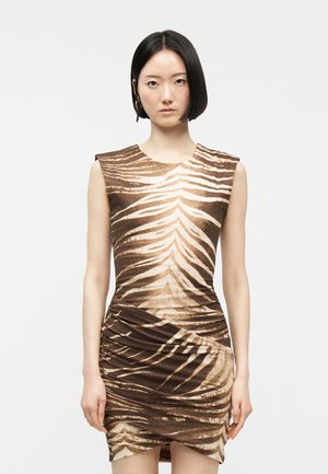 Lyhythiuksinen nainen, jolla on mustat hiukset ja joka on pukeutunut hihattomaan ruskeaan ja beigenväriseen eläinkuosiseseen mekkoon, seisoo yksinkertaisen vaalean taustan edessä.