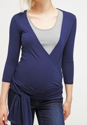 Zwangere vrouw draagt een marineblauw wikkelshirt over een grijs hemdje, gecombineerd met donkerblauwe spijkerbroek, staand tegen een effen achtergrond.