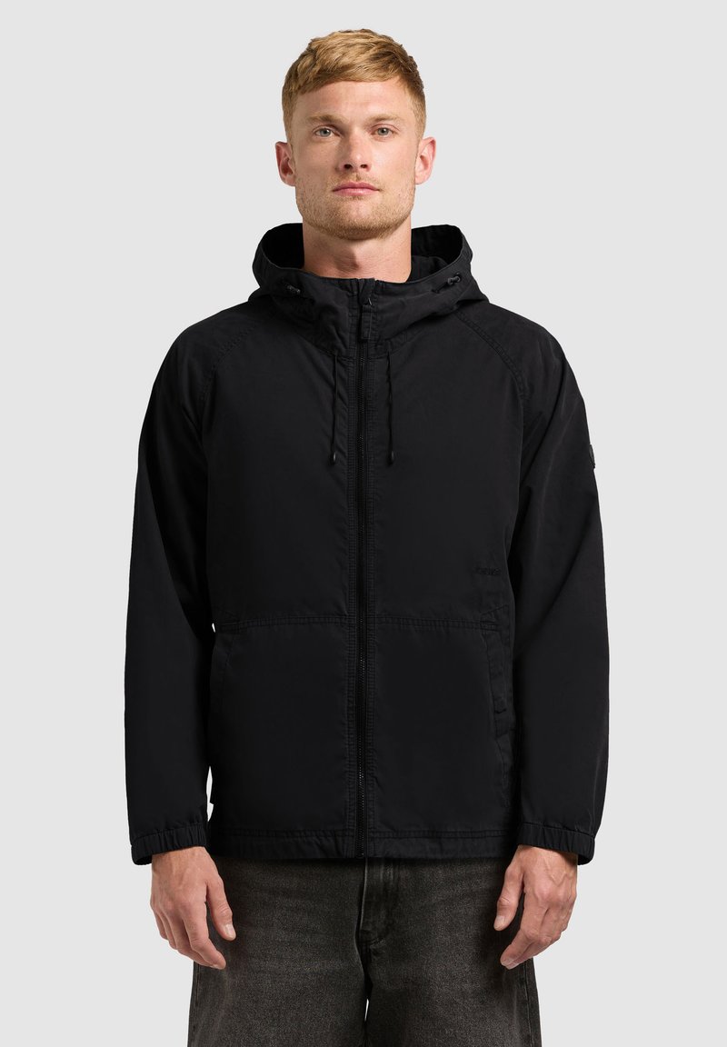 Homme aux cheveux blonds courts portant une veste à capuche noire et un jean gris foncé, debout devant un fond uni gris clair.