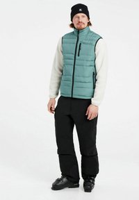 Poduszka w kolorze teal kamizelka z wysokim kołnierzem, czarnymi suwakami i beżowymi, teksturowanymi rękawami, w połączeniu z czarnymi spodniami i butami narciarskimi. Minimalne logo widoczne.