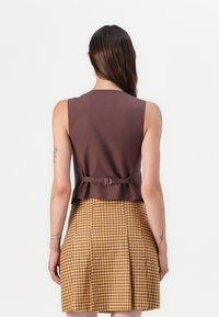 Top sans manches marron avec une silhouette ajustée et un détail de boucle au dos, associé à une jupe plissée à carreaux jaunes et marron.