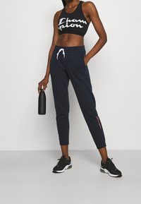 Svart sport-BH med vitt "Champion"-logotyp, kombinerad med marinblå joggingbyxor med vit dragsko och rosa logotypdetalj, bärs med svarta sneakers.