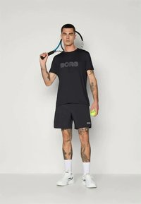 Μαύρο σπορτίφ T-shirt και σορτσάκι με λογότυπο "BORG", κρατώντας μια ρακέτα τένις και μια μπάλα. Αθλητική εφαρμογή, το ύφασμα φαίνεται ελαφρύ και αναπνεύσιμο.