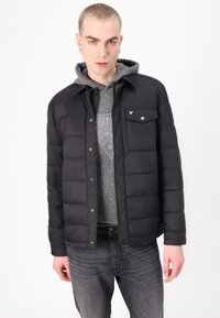 Chaqueta de plumón negra con un diseño acolchado, botones a presión plateados y un bolsillo en el pecho. Sudadera gris por debajo con un patrón texturizado.