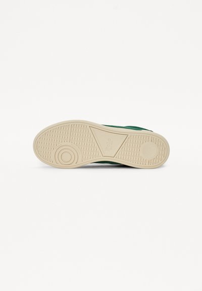 Adidași verzi cu un upper din piele de căprioară texturată, talpă din cauciuc crem și un accent circular din cauciuc pe partea de jos. Prezintă un model de tălpi în formă de triunghi.