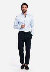 Lichtblauwe langärmige shirt met een knoopsluiting bij de kraag, donkerblauwe op maat gemaakte broek, en zwarte instapschoenen, tegen een effen achtergrond.