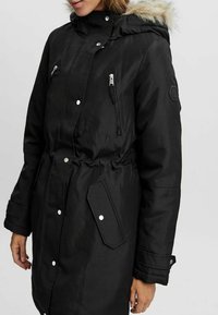 Parka noire avec une capuche en fourrure, deux poches poitrine zippées, une fermeture à boutons-pression et une taille réglable. Tissu lisse et texturé.