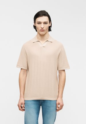 Joven con cabello oscuro que lleva una camisa polo beige de manga corta con sutiles patrones verticales y jeans azul claro, de pie frente a un fondo blanco.