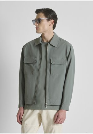 Man draagt een grijze jas met rits en klepzakken, lichtgroen shirt, beige broek en donkere zonnebril, kijkt naar links tegen een grijze achtergrond.