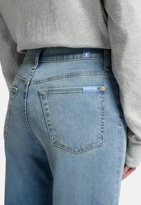 Light blue denim jeans with a high waist, featuring back pockets and subtle distressing. Accented by a small blue tag on the waistband.