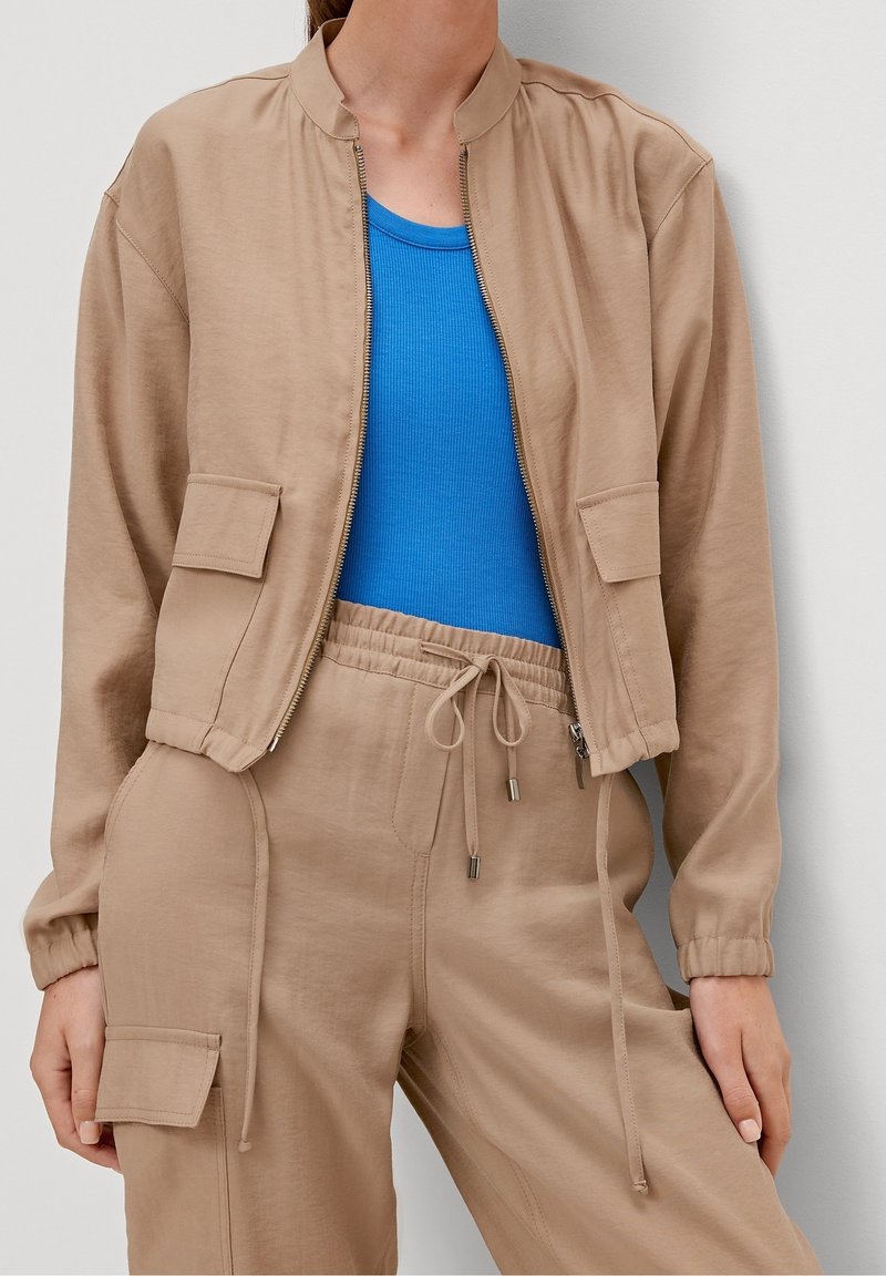 Beige bomberjakke med lynlås foran, ribbet krave og to frontlommer, parret med en blå ribbet tanktop og matchende beige bukser.