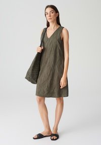 Khaki green sleeveless dress with a V-neck, featuring a textured leaf pattern, paired with a woven black tote bag and black slide sandals.