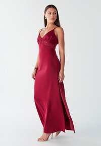 Bordinės spalvos satino maxi suknelė su nėrinių įsiuvimu liemens srityje, giliais V formos iškirpte, plonomis petnešėlėmis ir šoniniu plyšiu.