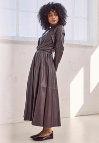 Robe en cuir brun foncé, longue avec des manches longues, taille cintrée et jupe évasée. Associée à des ballerines noires, sur fond clair.