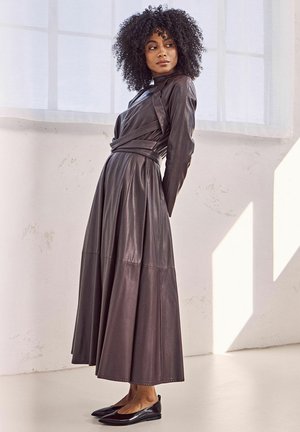 Long, dark brown leather dress with long sleeves, cinched waist, and flared skirt. Paired with black flats, set against a light background.