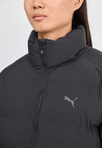 Melnā pūkaina jaka ar augstu apkakli un priekšējo rāvējslēdzi. Uz krūtīm ir smalks pelēks Puma logo. Gluds, izolēts audums.
