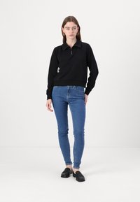 Fekete pulóver, cipzáras gallérral, bordás mandzsettákkal és derékrésszel, kék skinny farmerral és fekete loaferekkel viselve. Egyszerű dizájn.