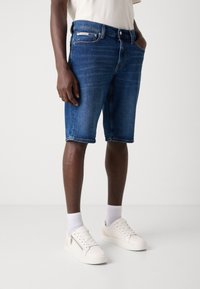 Tamne traperice u tamnoplavoj boji s klasičnim krojem, pet džepova, kontrastnim šavovima i oznakom Calvin Klein na pojasu.