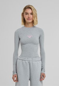 Lys grå langermet topp med tettsittende design, med et rosa sommerfuglmotiv i midten. Mykt bomullsmateriale med en jevn tekstur.